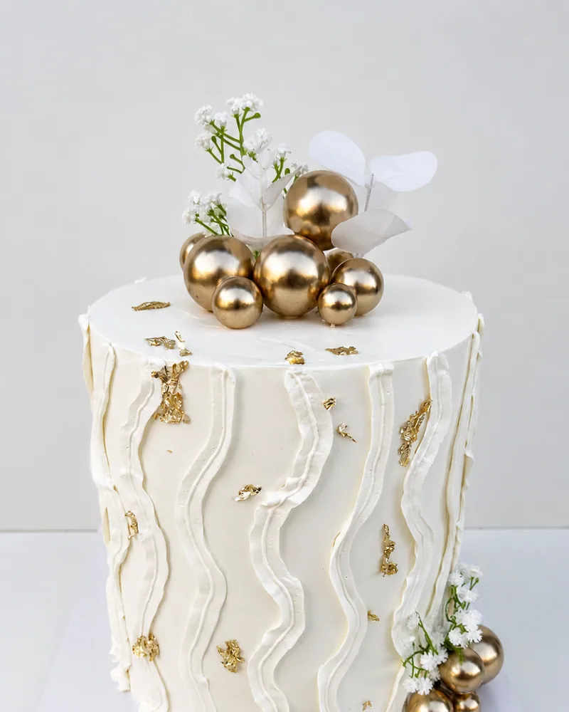 A single-tier white celebration cake with a soft wave texture, gold spheres and baby's breath, by The Cake Studio Harare