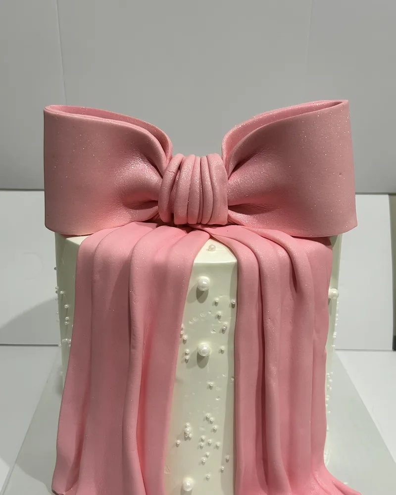 A tall ivory cake wrapped in a large pink fondant bow and draped sash with white pearls, by The Cake Studio Harare