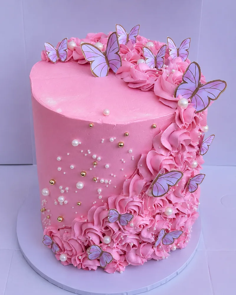 A pink buttercream cake with piped ruffles, pearls, gold dragees and lilac butterflies, by The Cake Studio Harare