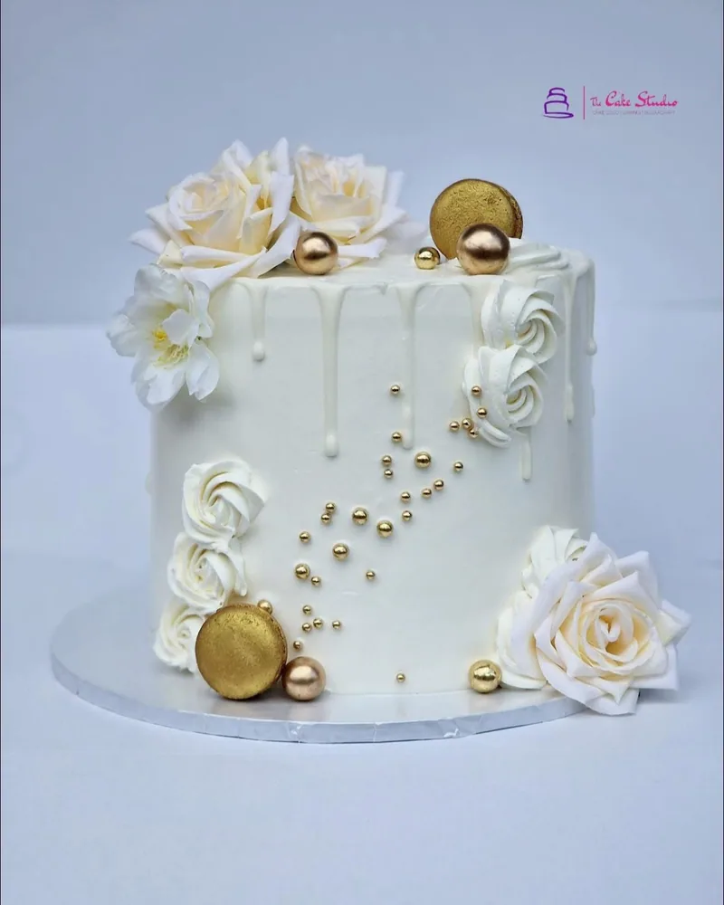 A single-tier white and gold drip cake with gold macarons and pearls, by The Cake Studio Harare